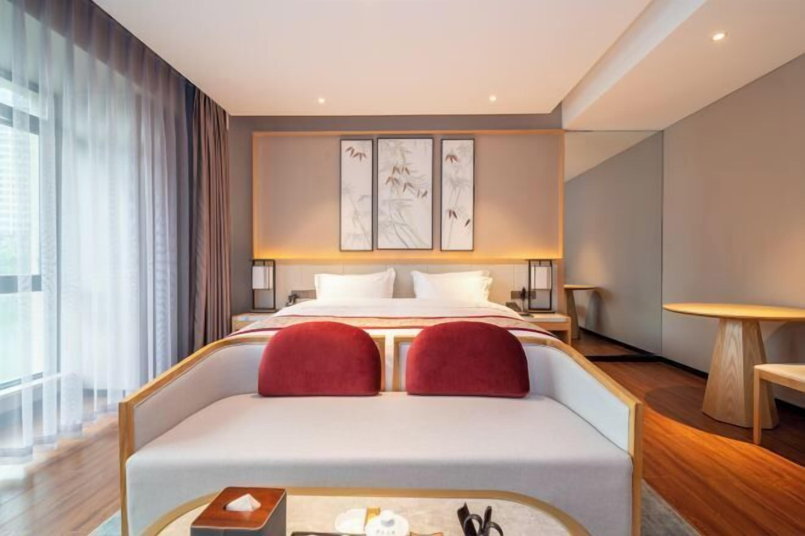 Una habitación de hotel moderna con una cama grande, un rincón de descanso con cojines rojos, una mesita con artículos de aseo y vistas a la ciudad a través de amplias ventanas.