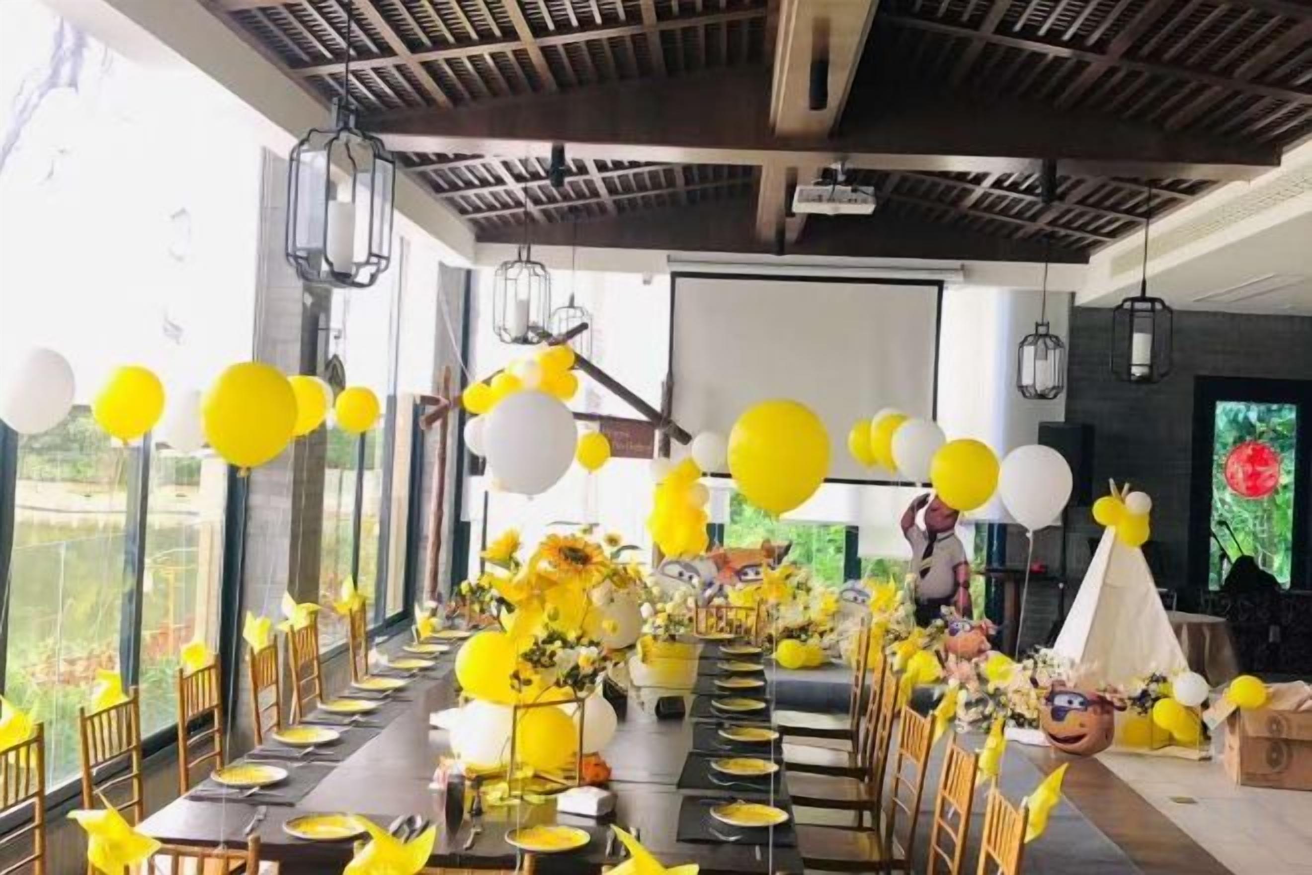 A long dining table set for a celebration with yellow and white balloons, floral centerpieces, and a projector screen in the background.