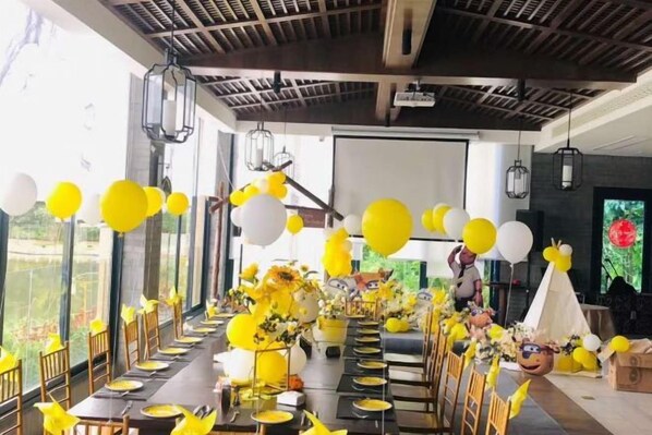 A long dining table set for a celebration with yellow and white balloons, floral centerpieces, and a projector screen in the background.