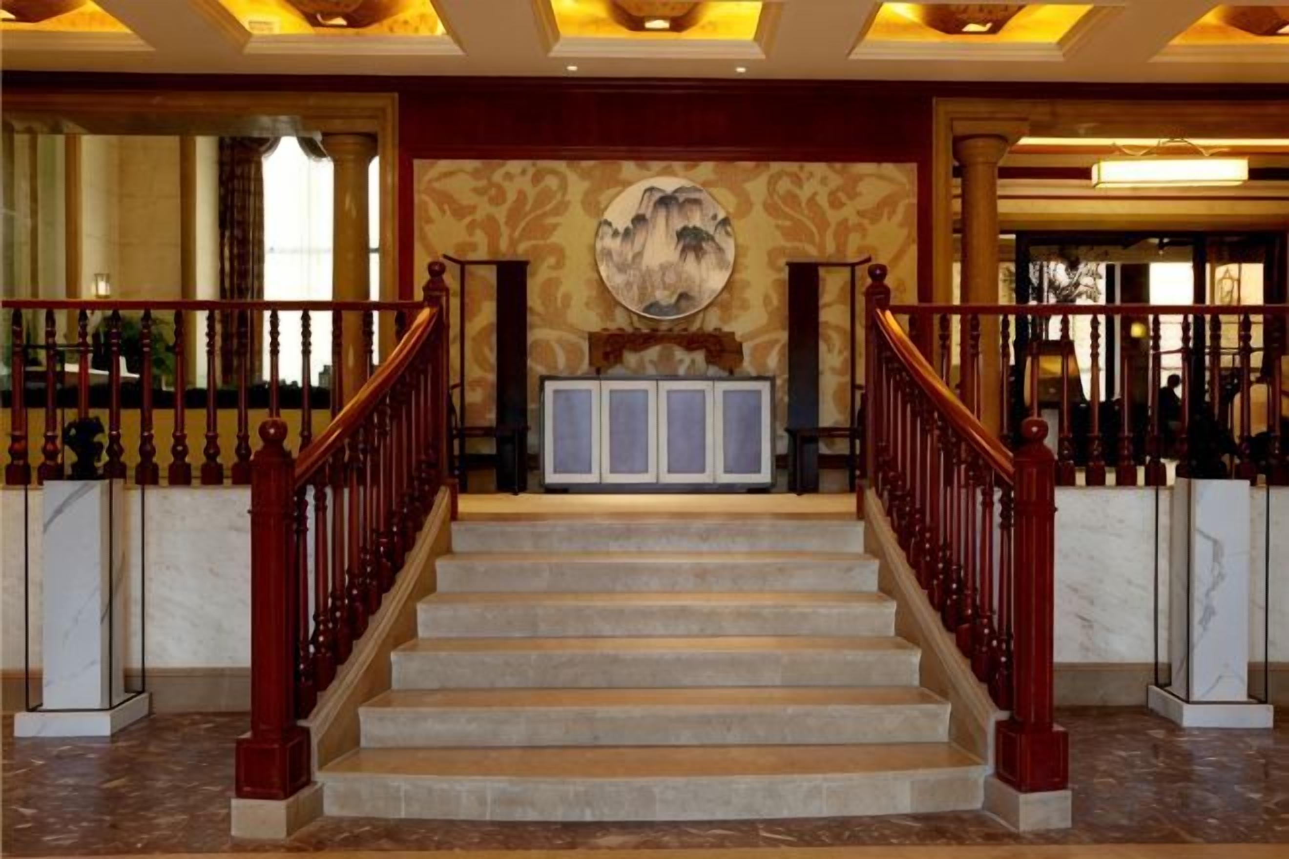 Una imponente escalera con escalones de mármol y barandas de madera, que conduce a un área de recepción con un mural grande y una obra de arte enmarcada sobre un mostrador.