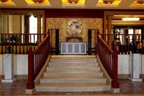 Una imponente escalera con escalones de mármol y barandas de madera, que conduce a un área de recepción con un mural grande y una obra de arte enmarcada sobre un mostrador.