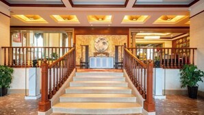 A grand staircase with wooden railings and a marble floor, leading to a lobby area with a decorative wall and a large circular artwork. - Aile Holiday Hotel (Sanming)