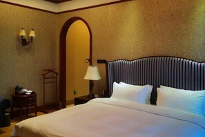 Habitación de hotel con una cama grande, cabecera de madera, armario, escritorio con lámpara y lámpara de pared.