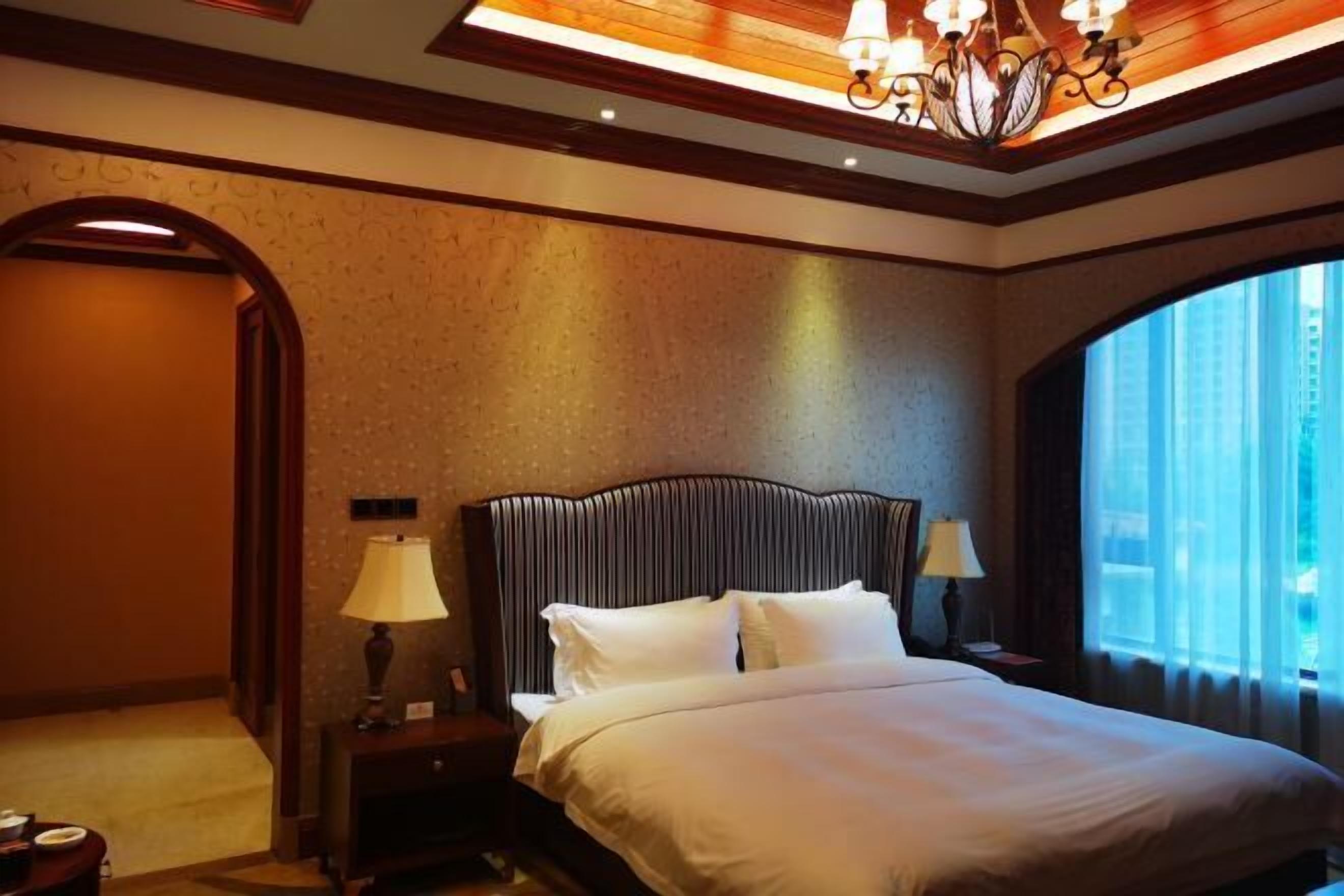 A hotel room with a large bed, two bedside tables with lamps, a chandelier, and a large window with curtains.