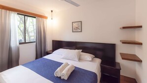 A neatly made bed with white and blue bedding, a ceiling fan, a bedside table, and a window with curtains.
