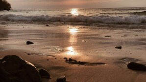 A beach scene with the sun setting over the horizon, waves gently lapping at the shore, and scattered rocks on the sand.