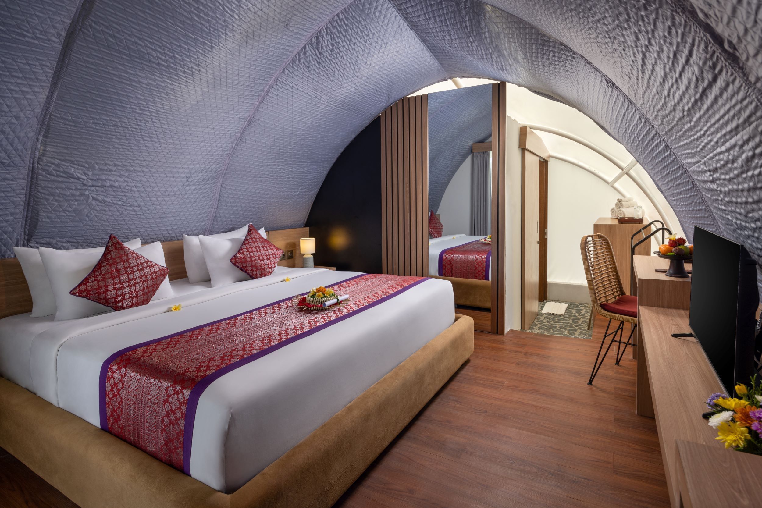 A bedroom with a curved ceiling, a bed with red and white bedding, a wooden bedside table, a chair, and a desk with a computer.
