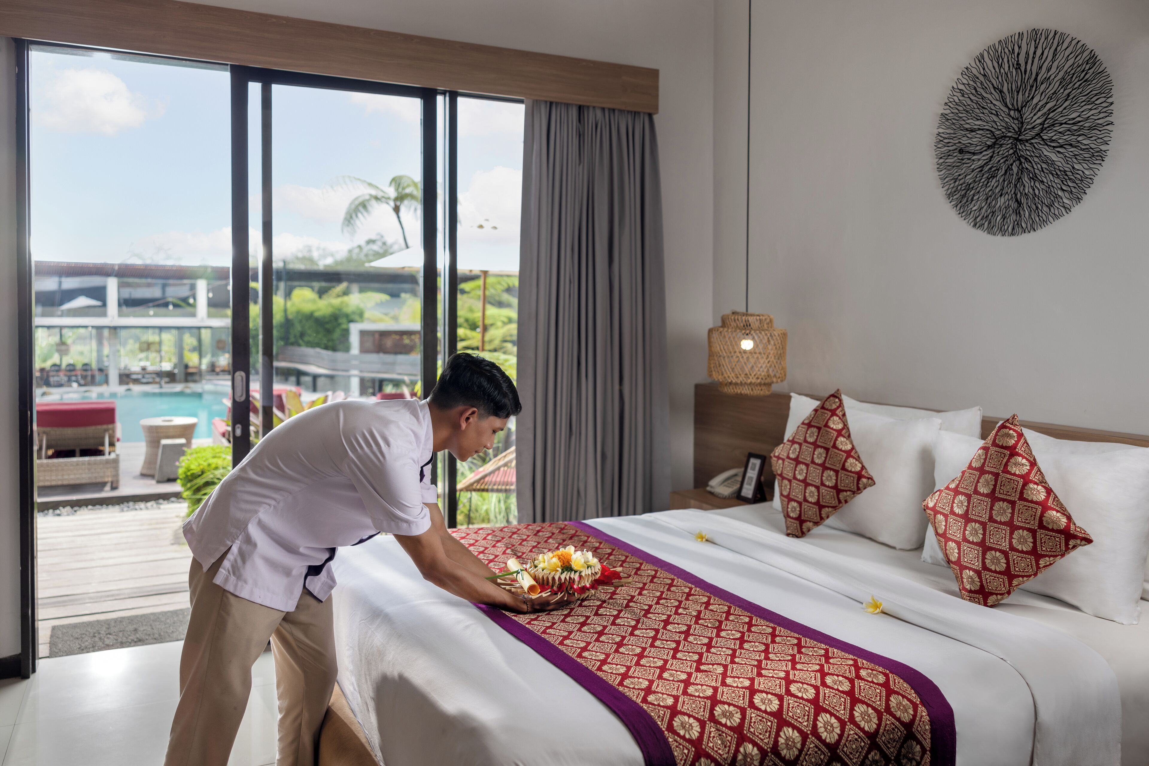 A hotel room with a bed, a bedside table, a lamp, and a view of a pool and greenery.