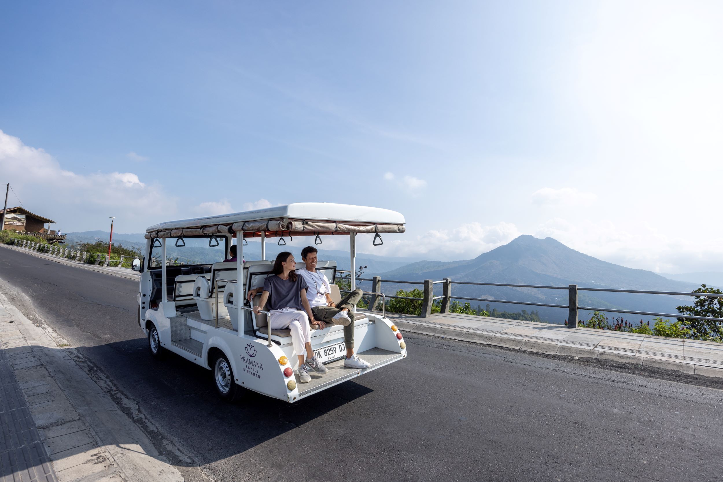 A white golf cart with two passengers on a paved road with a mountain in the background.