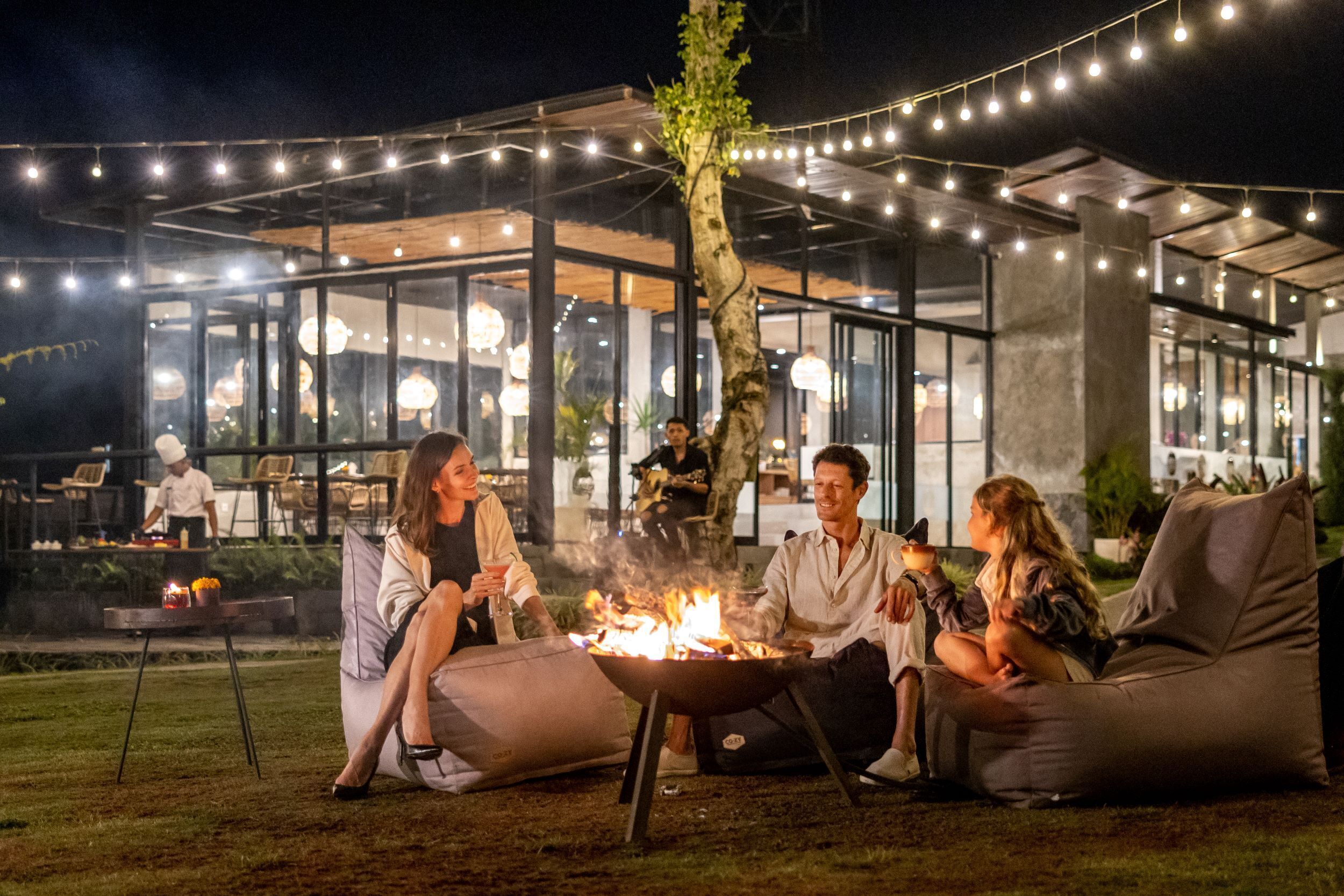 An outdoor seating area with a fire pit, surrounded by string lights and modern architecture.