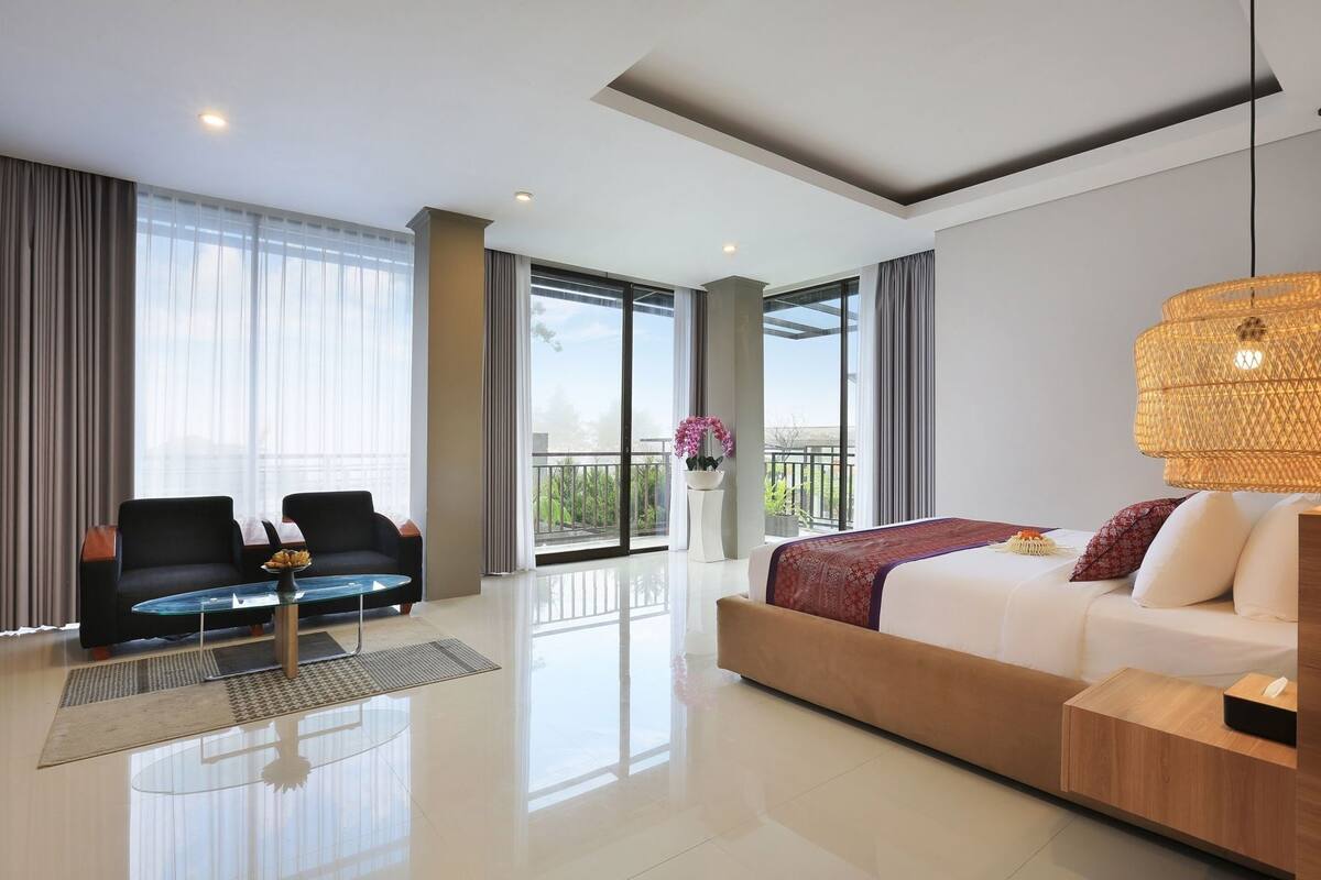 Une chambre d’hôtel moderne, dotée d’un grand lit, de deux fauteuils, d’une table basse en verre et d’un balcon avec vue.
