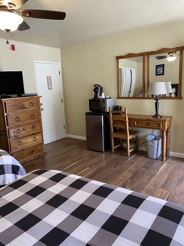 Habitación de hotel con cama, cómoda de madera, escritorio con silla, microondas, refrigerador y un cesto de basura.