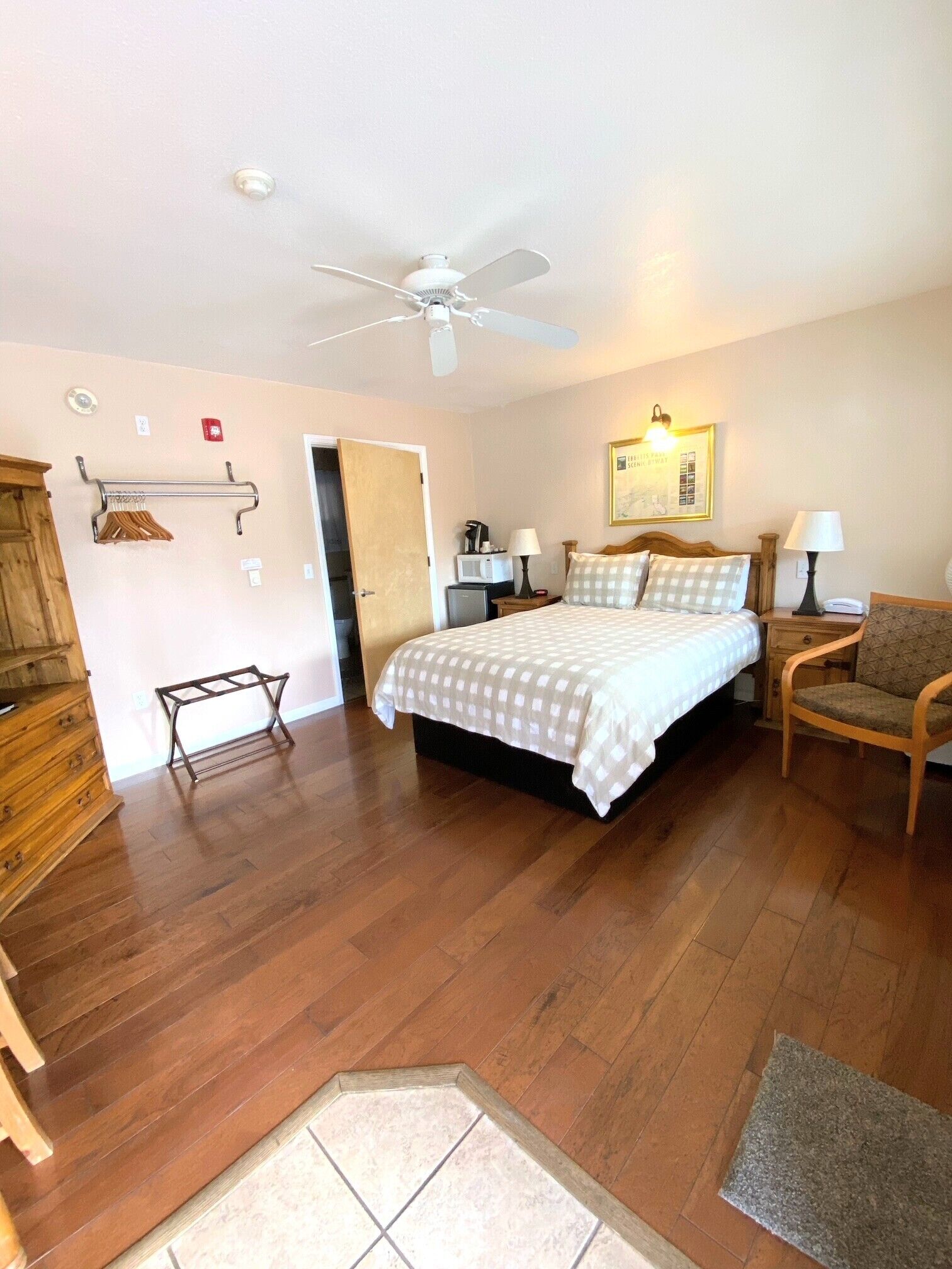 A bedroom with a bed, a ceiling fan, a wooden dresser, a chair, a small table, and a microwave.