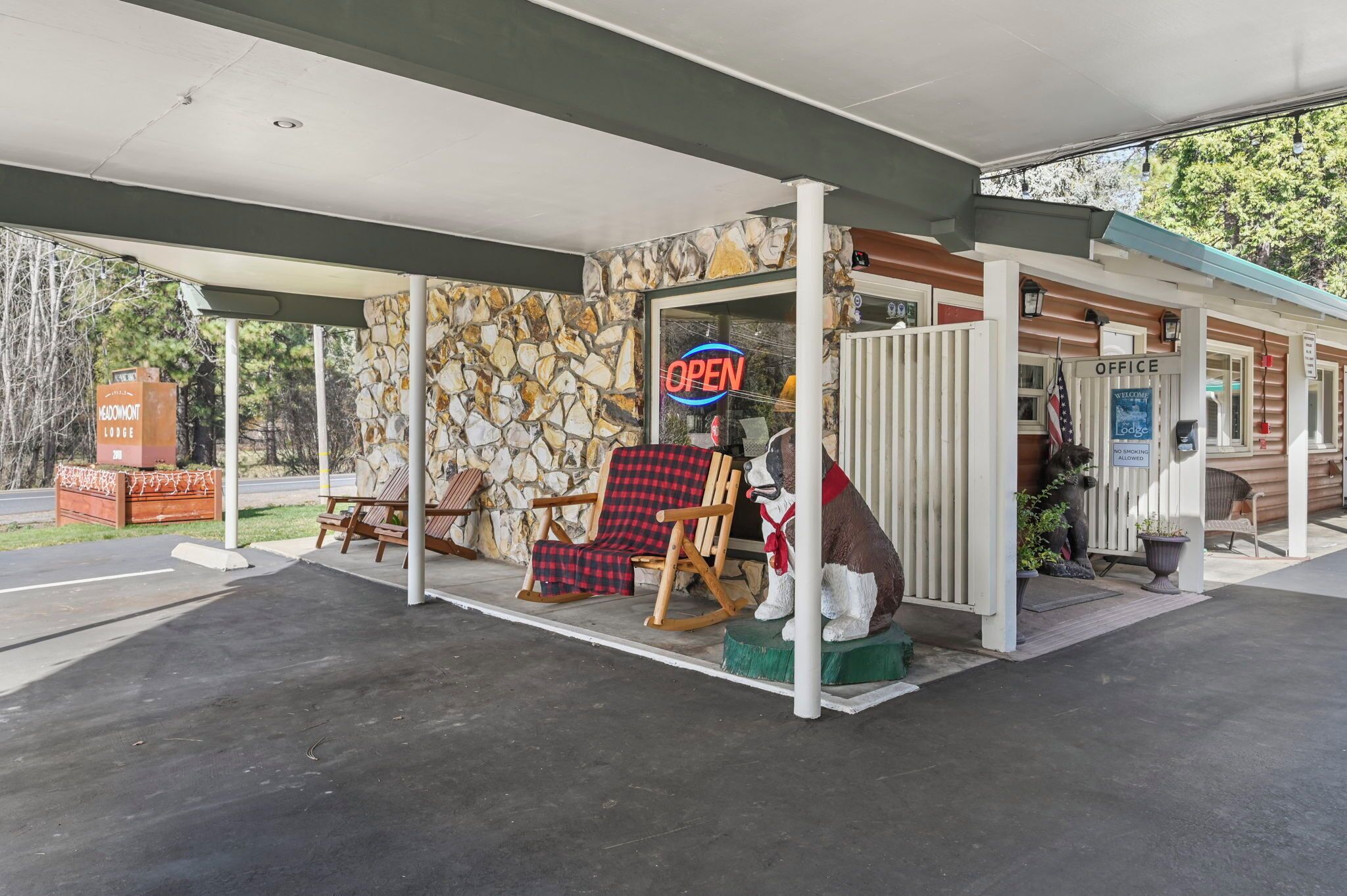 Un motel à la façade en pierre, avec une enseigne « OUVERT », une chaise à bascule en bois, un fauteuil à carreaux rouges et une statue de chien.