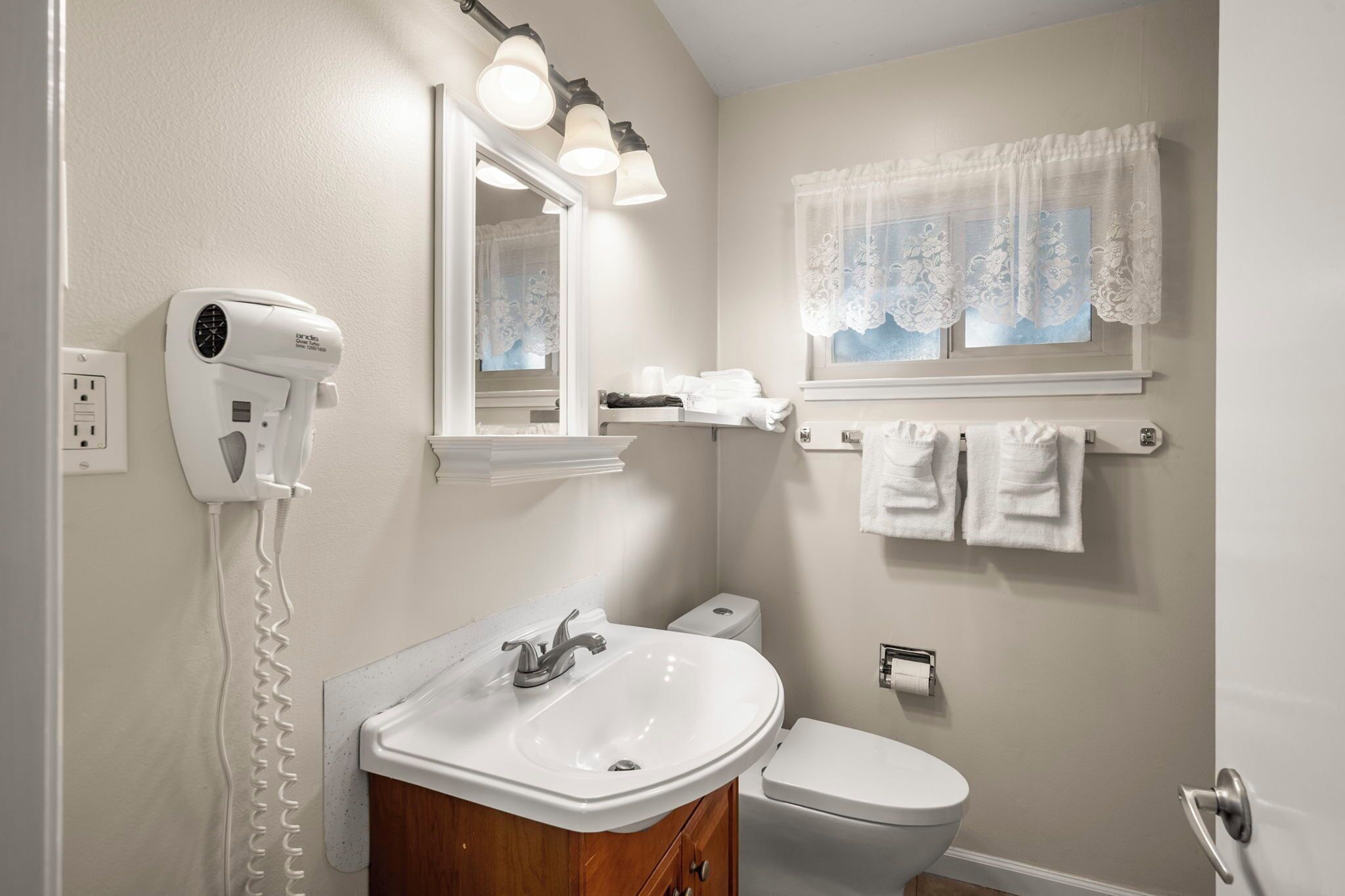 Een badkamer met een witte wastafel, toilet, haardroger bevestigd aan de muur en een raam met kantgordijnen.