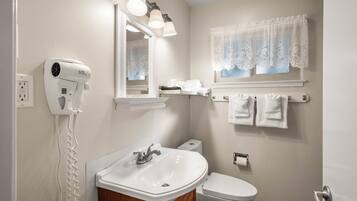 Une salle de bain avec un lavabo blanc, des toilettes, un sèche-cheveux fixé au mur et une fenêtre ornée de rideaux en dentelle.