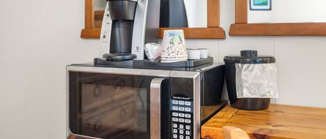 Un microondas con un café preparador encima, un pequeño cubo de basura y una mesa de madera.