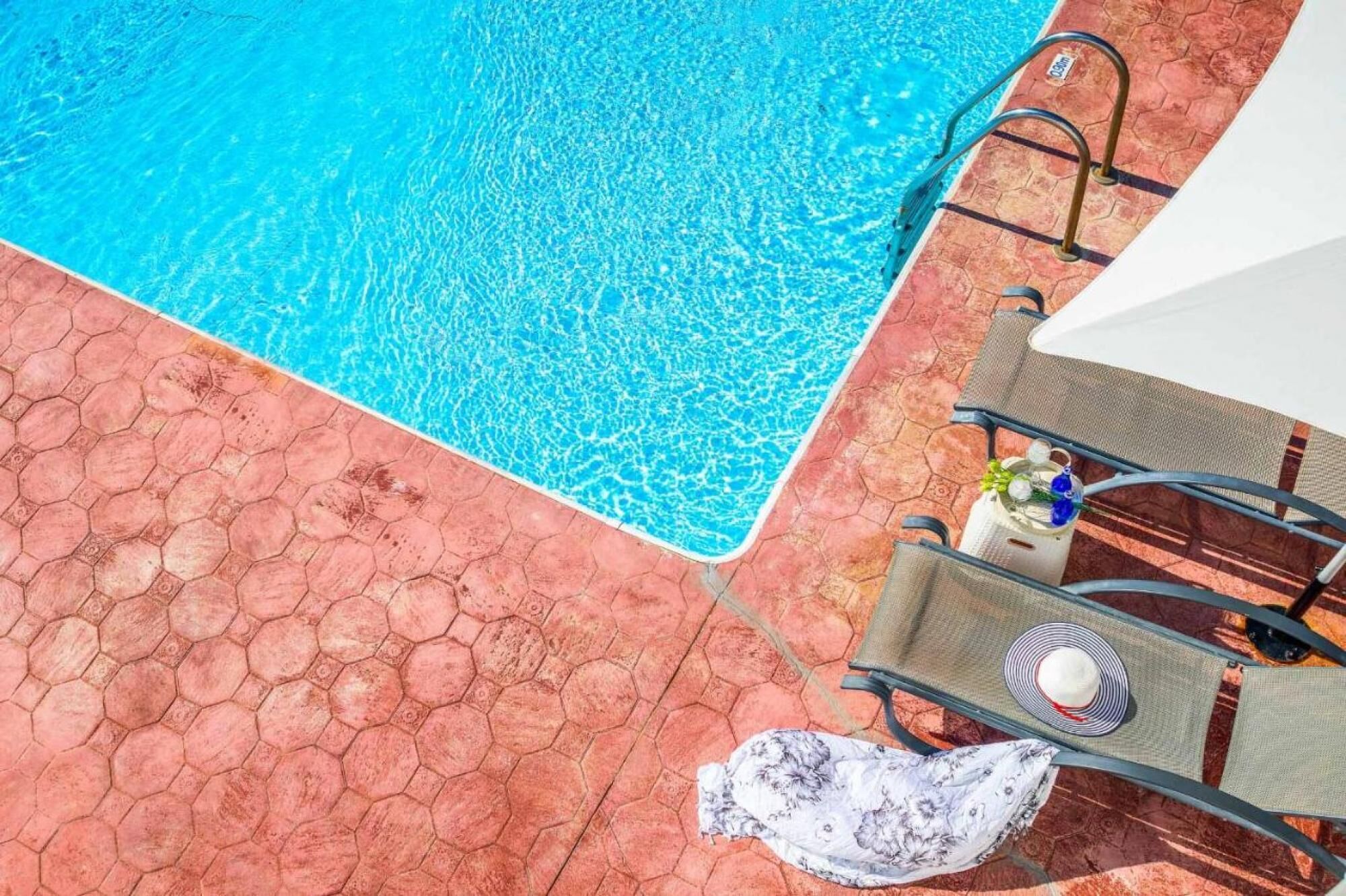 A pool area with a hexagonal tiled floor, a pool ladder, and lounge chairs with a table and a decorative item.