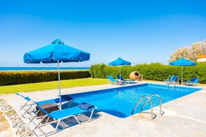 Piscina com espreguiçadeiras azuis e guarda-sóis, rodeada por um arbusto e vista para o mar.
