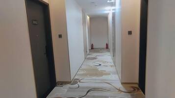 A hotel corridor with a patterned carpet, doors on either side, and a fire extinguisher in the distance.