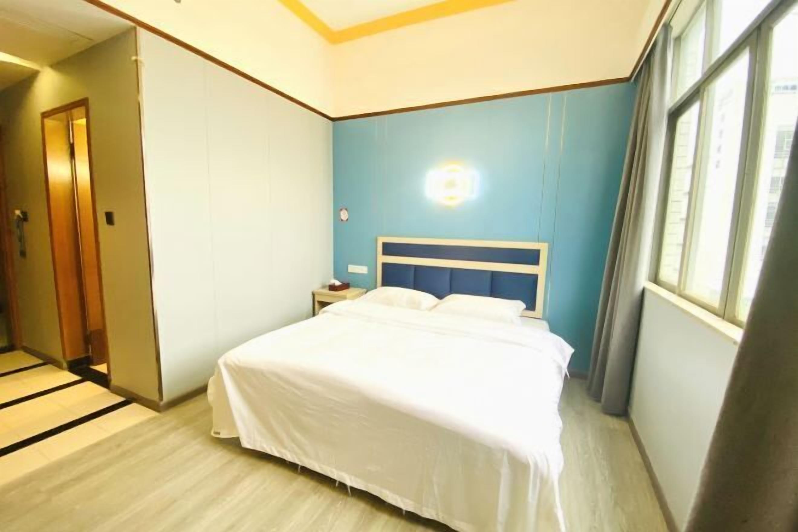 Una habitación de hotel con cama, mesita de noche, lámpara de pared y ventana con cortinas.