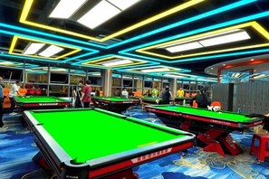 Una sala de billar con múltiples mesas de billar, iluminación colgante y una alfombra estampada.