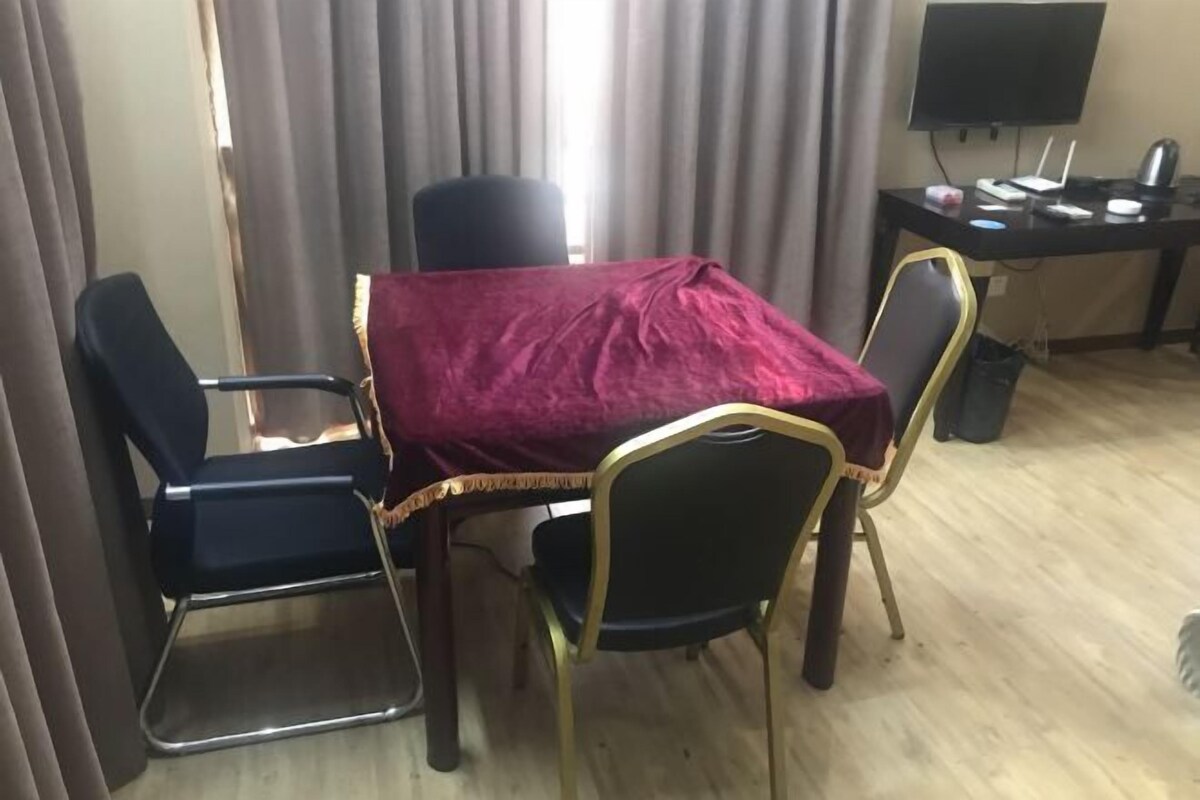 A room with a table covered by a red cloth, four chairs, a desk with a computer, and a TV.