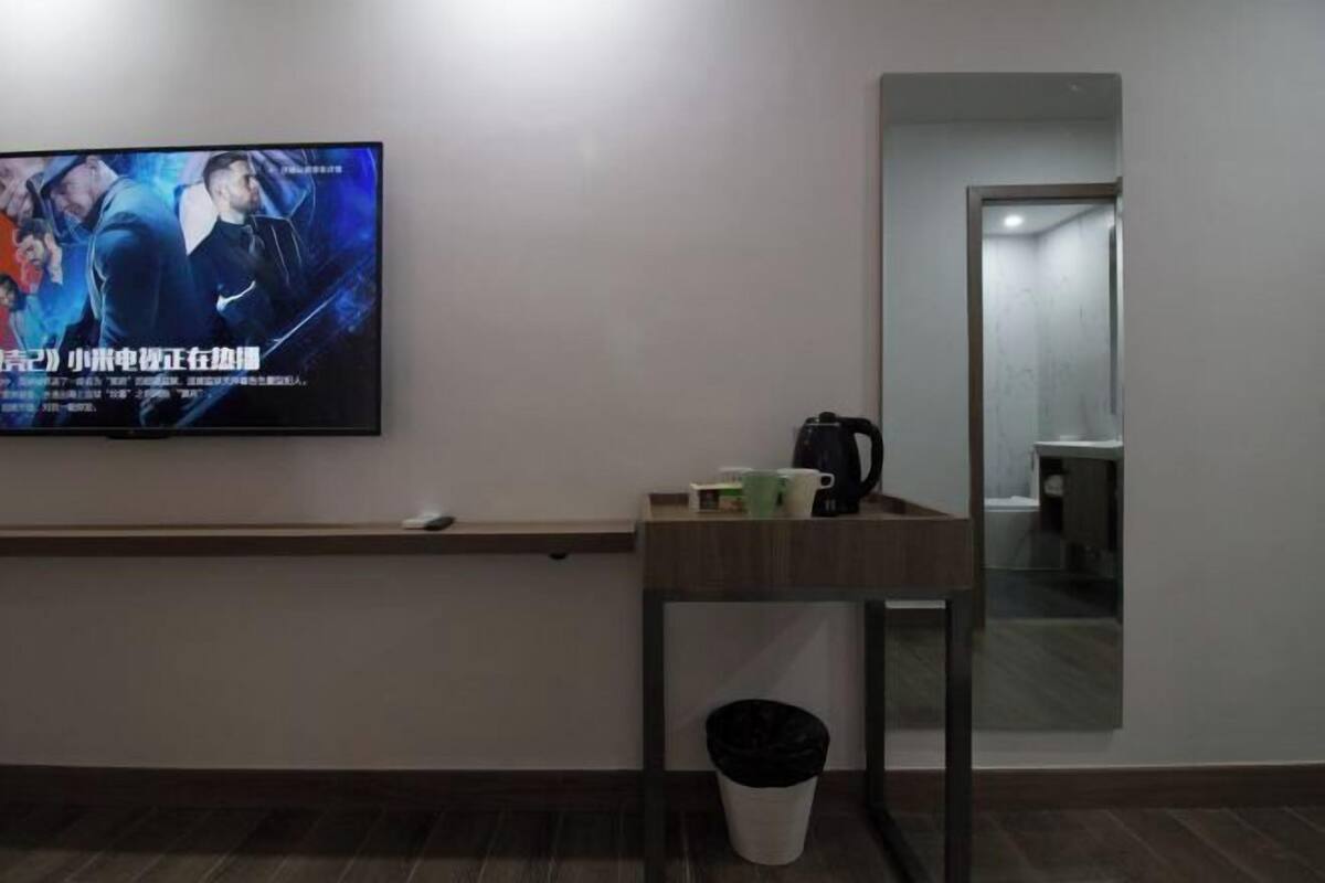 Une chambre d’hôtel avec un téléviseur fixé au mur qui diffuse une affiche de film, une petite table avec une machine à café et des tasses, une poubelle et un miroir au mur.