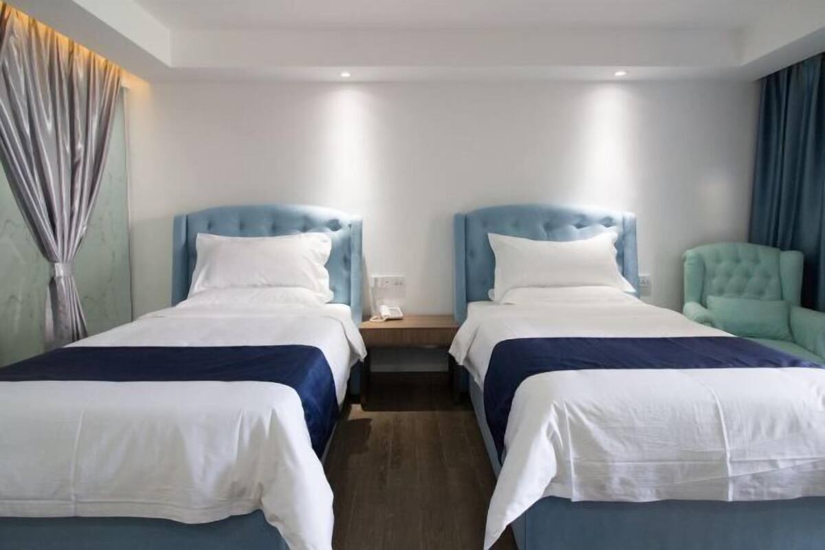 Une chambre d’hôtel avec deux lits, chacun doté d’une tête de lit bleue et de draps blancs. On y trouve une petite table de chevet avec une lampe et un téléphone.