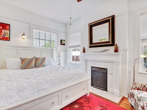 Quarto com cama branca, lareira e janela com persianas.