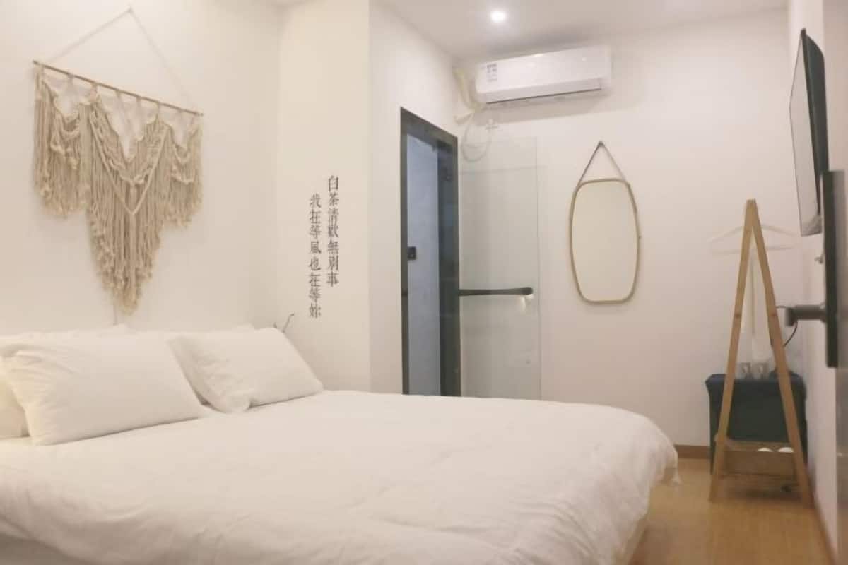 A bedroom with a bed, a wall-mounted macrame piece, a door, a mirror, and a standing fan.