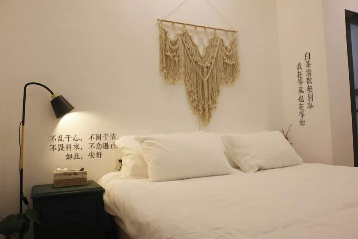 A neatly made bed with white bedding and pillows, a bedside lamp, and a wall-mounted macrame piece.