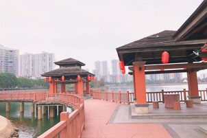 A traditional pavilion with a wooden roof and red lanterns, situated by a calm body of water with a bridge leading to it.