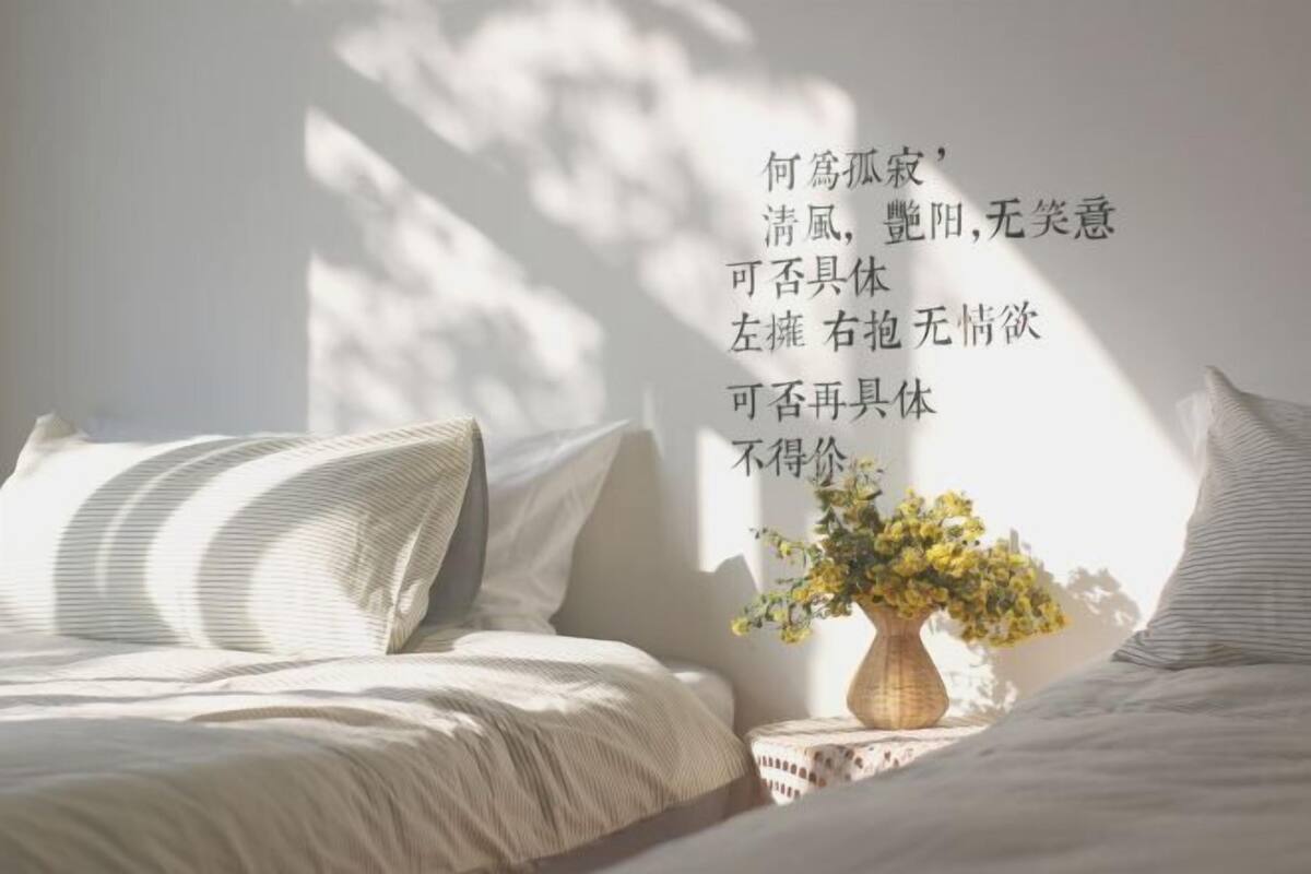 A bedroom with a bed, pillows, a bedside table with a vase of flowers, and a wall with text.