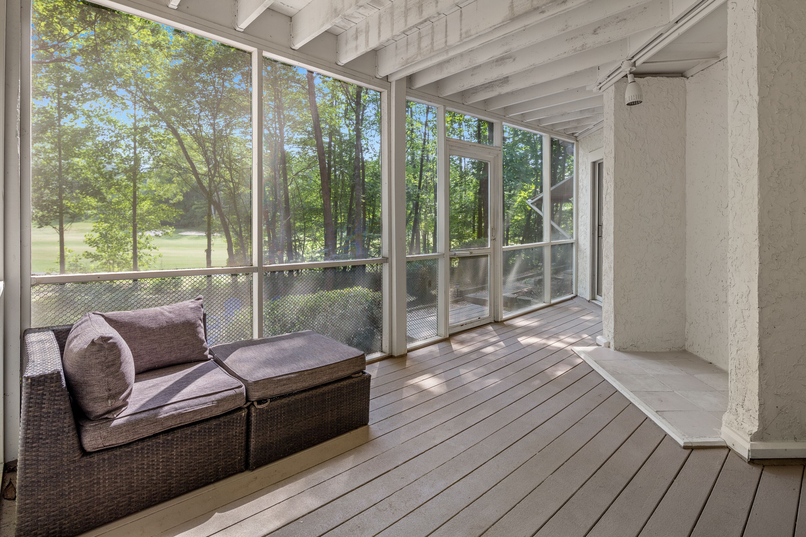 A covered porch with a wicker sofa and large windows offering a view of a wooded area.