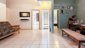 A spacious living area with a patterned sofa, a bench, a dining table set for two, and a hallway leading to another room.
