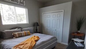 Un dormitorio con una cama, un ventilador de techo, una ventana con persianas, una puerta, una mesita de noche con una lámpara y un cartel en la pared.