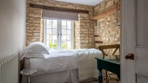 Un dormitorio con pared de piedra, ventana con vistas, cama con ropa blanca y una silla de madera.