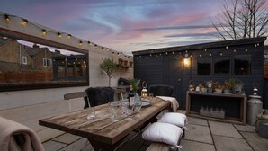 Un patio al aire libre con mesa de madera dispuesta para comer, rodeado de cojines y plantas en macetas.