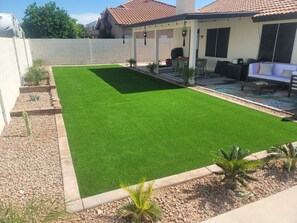 Een achtertuin met kunstgras, een terrasgedeelte met tuinmeubilair, en een witte omheining.
