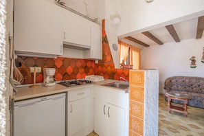 A compact kitchen with white cabinets, a gas stove, and a coffee maker. A small wooden stool is near the sink, and a patterned sofa is in the adjacent living area. - Holiday Home Vistamar 4 with Sea Views, Garden and Wi-Fi (Vinaròs)
