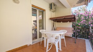 Un balcón con mesa y sillas, una puerta corrediza de vidrio, un equipo de aire acondicionado y un arbusto de flores.