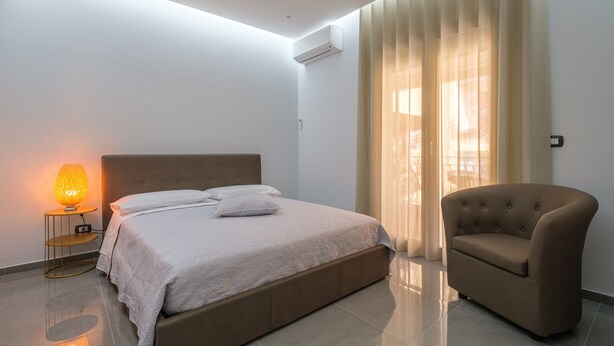 Un dormitorio con cama, lámpara de noche, silla y cortinas.