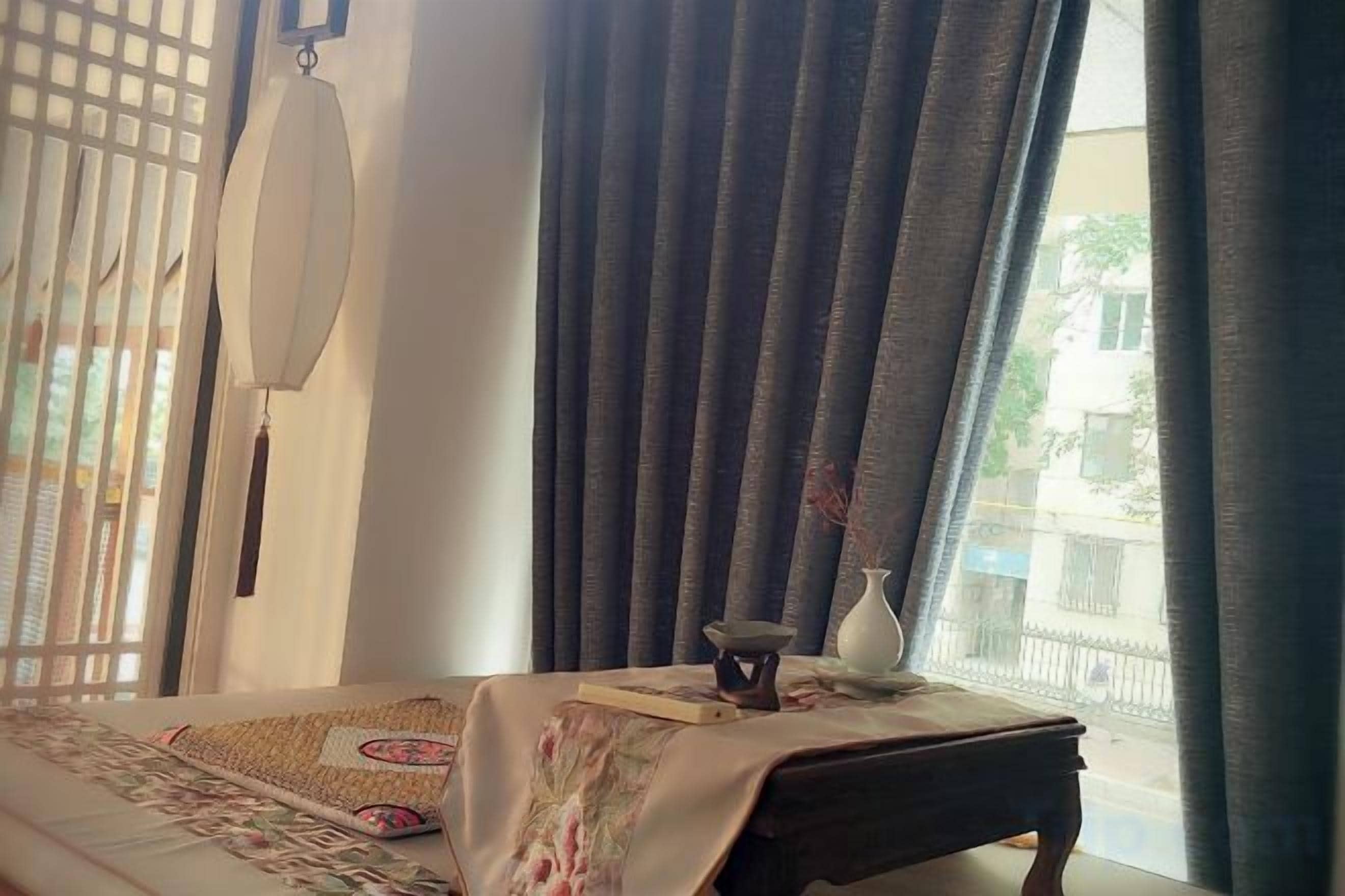 Une chambre avec un lit, une petite table sur laquelle se trouve un vase, et une vue par la fenêtre, qui est ornée de rideaux.