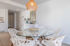 Uma área de refeições com mesa de vidro, cadeiras brancas, luminária pendente de vime e um armário branco com objetos decorativos.