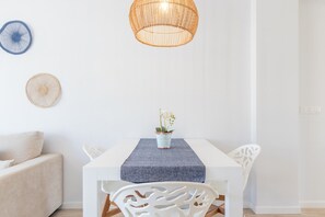 Uma área de refeições com uma mesa branca, um vaso azul estampado com uma orquídea e uma luminária pendente de rattan trançado.