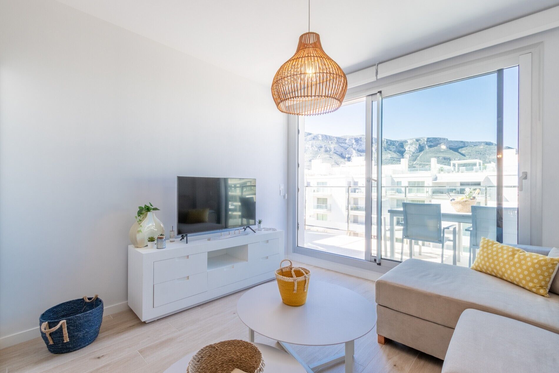 A modern living room with a TV, sofa, and a balcony with a view of buildings and mountains.