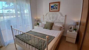 Un dormitorio con cama, mesita de noche, lámpara, cuadro enmarco y ventana con cortinas translúcidas.