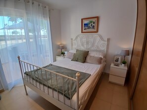 Une chambre à coucher avec un lit, une table de chevet, une lampe, un tableau encadré et une fenêtre avec des rideaux légers.