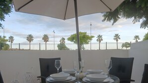A table set for two with white plates, glasses, and a bowl, under a white umbrella, with chairs around it.